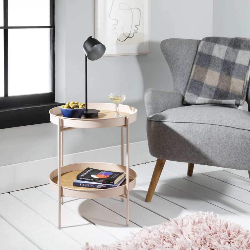 A blush pink, two-tier round side table with rose gold accents, placed next to a gray armchair with a lamp and decorative items on its top tier.