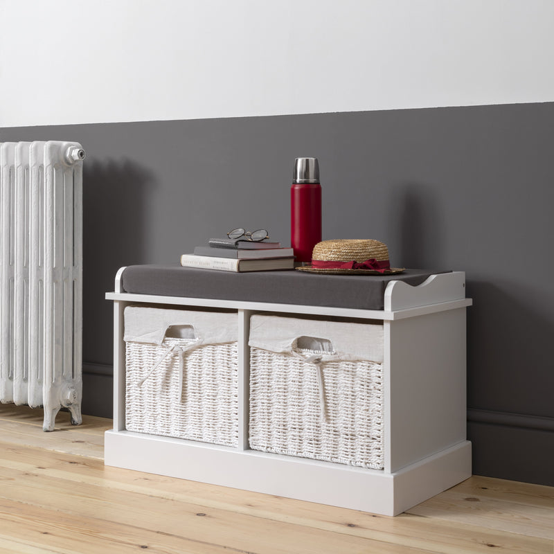 A white storage bench with two baskets placed on the floor beside a radiator, with a gray wall in the background.