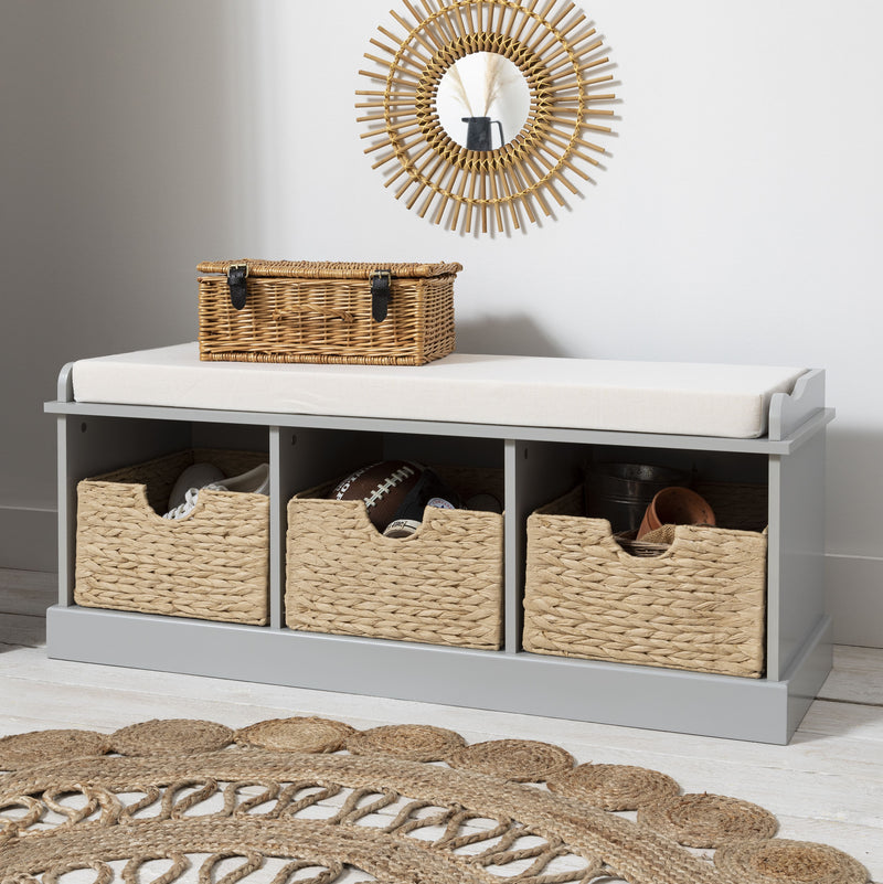 A gray storage bench with three brown wicker baskets, placed on a white rug with a decorative sun-like mirror above it.