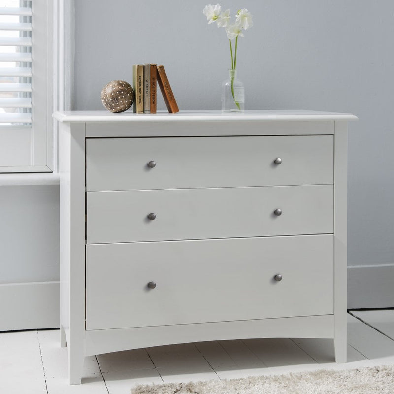 A classic white chest of drawers with bronze handles, situated in a room with a book, a vase with flowers, and a rug.